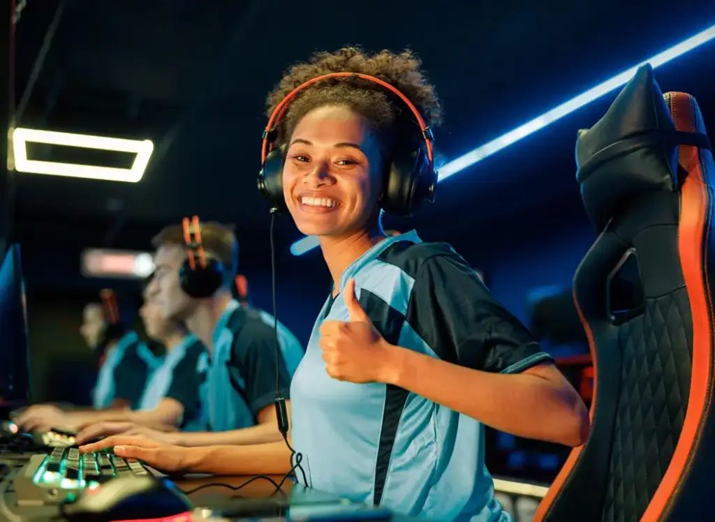 Een vrouwelijke esports-speler draagt een headset en glimlacht terwijl ze achter haar gamingcomputer zit. De sfeer in de gamingruimte is kleurrijk en energiek.